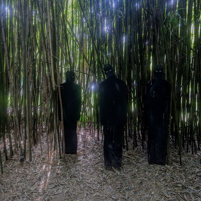Three eerie, shadowy human-shaped wooden figures with glowing eyes stand in a dense bamboo grove with sunlight filtering through the stalks.Three eerie, shadowy human-shaped wooden figures with glowing eyes stand in a dense bamboo grove with sunlight filtering through the stalks.